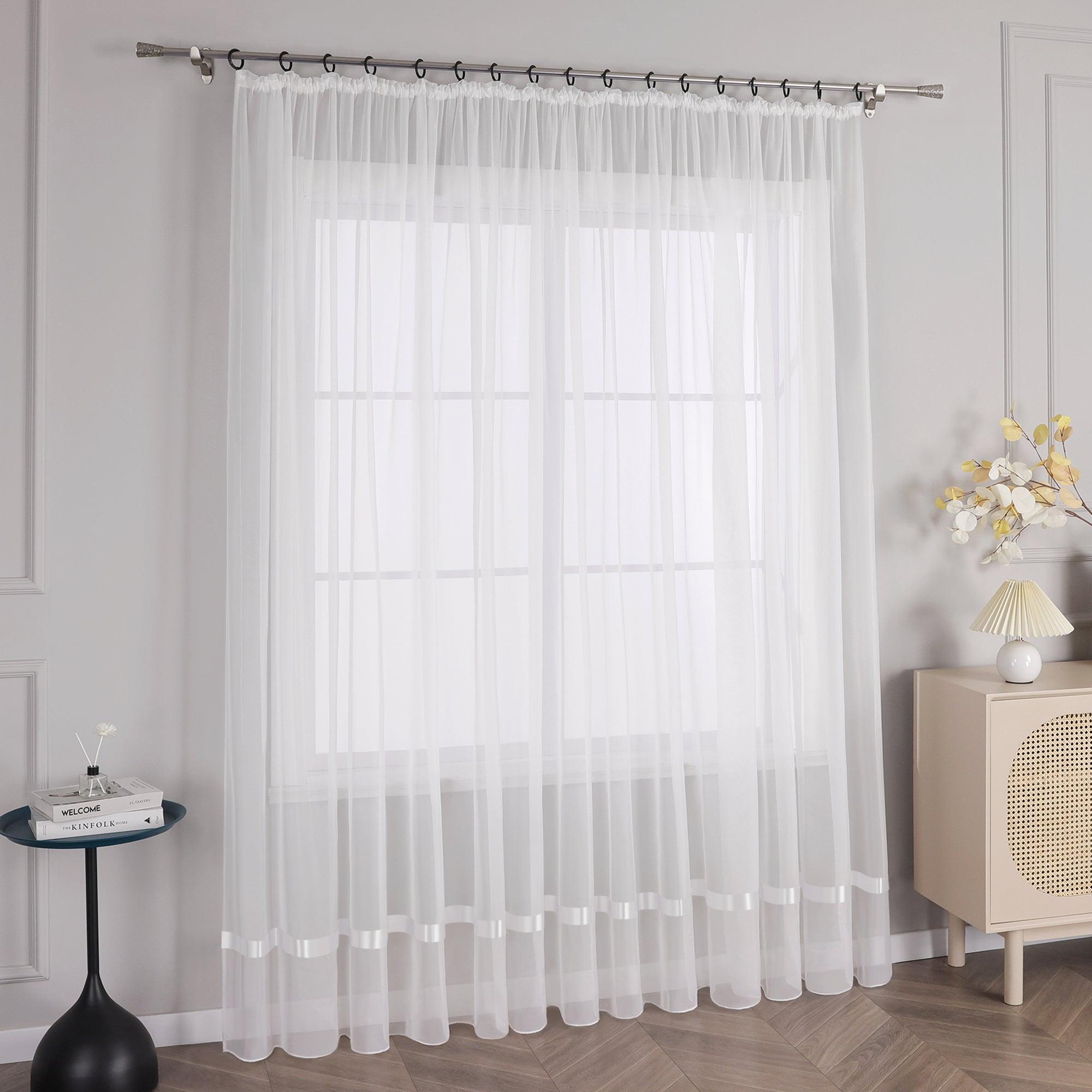 Vor der hellgrauen Wand befindet sich ein Fenster mit weißen Voile-Vorhängen mit Metallstangen mit schwarzen Ringen am oberen Ende. Links vom Fenster steht ein kleiner schwarzer runder Tisch mit ein paar Büchern und Vasen mit Blumen darauf. Die Vorhänge sind lang und hängen bis zum Boden, sie sind weiß und lichtdurchlässig.