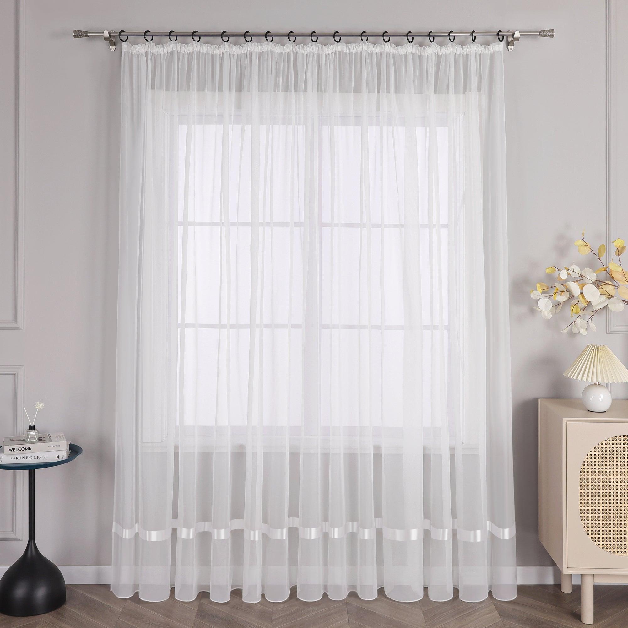 An einem Fenster hängt ein Vorhang mit Kräuseln, und am oberen Ende des Vorhangs befindet sich eine Metallstange mit schwarzen Ringen, die den Vorhang an seinem Platz halten. Auf der linken Seite des Fensters steht ein kleiner schwarzer runder Tisch, auf dem einige Gegenstände stehen. Rechts vom Fenster steht ein heller Schrank mit einer Lampe mit hellem Schirm und einem Strauß dekorativer Blumen.