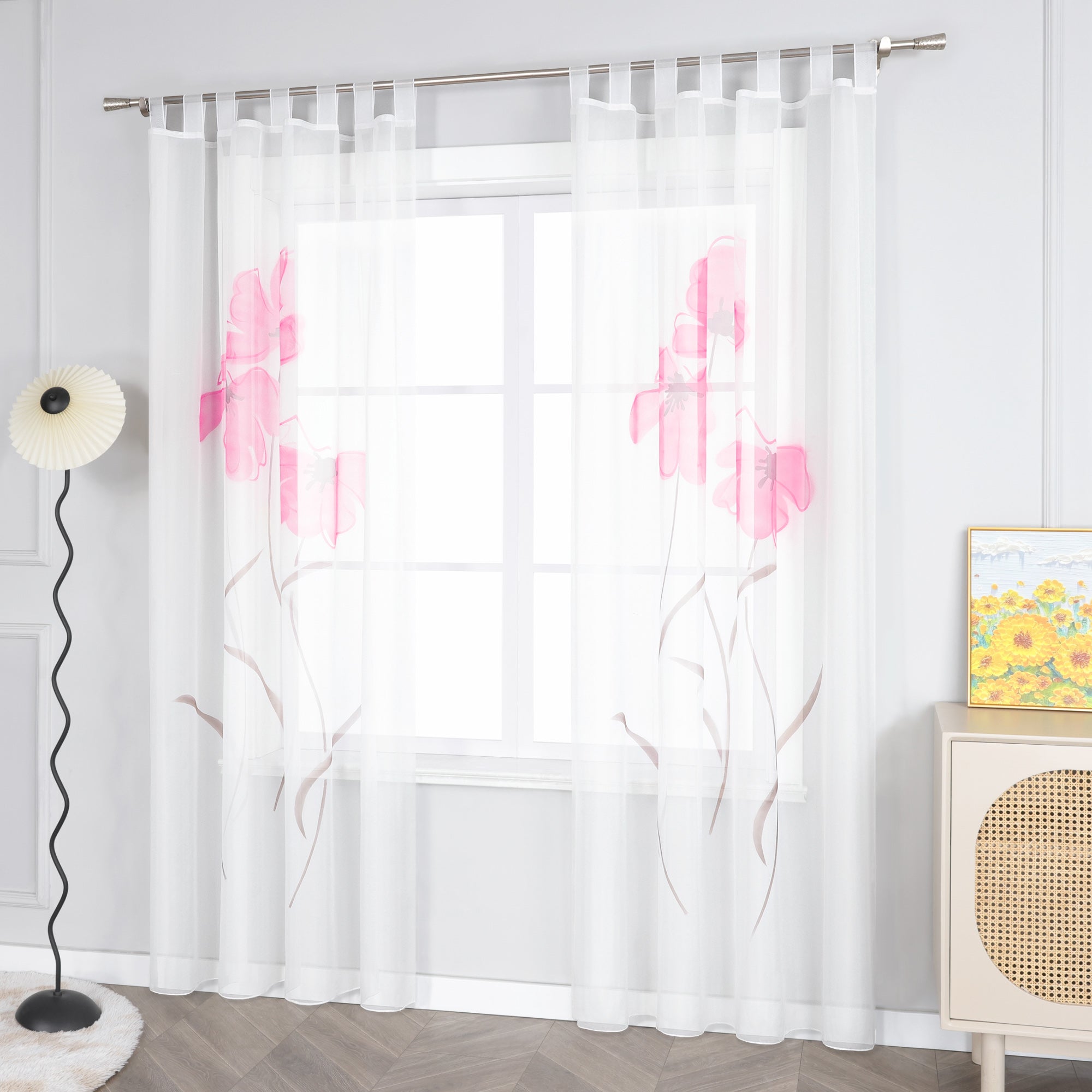 Das Bild zeigt einen weißen Voile-Vorhang mit einem Muster aus rosa Blumen und braunen Zweigen, der mit Metallringen an einer Stange aufgehängt ist. Neben dem Fenster steht eine Stehlampe mit einem weißen Lampenschirm. Daneben befindet sich ein heller Schrank mit einem gewobenen Musterelement auf der Oberfläche, auf dem ein Gemälde in Gelbtönen platziert ist.