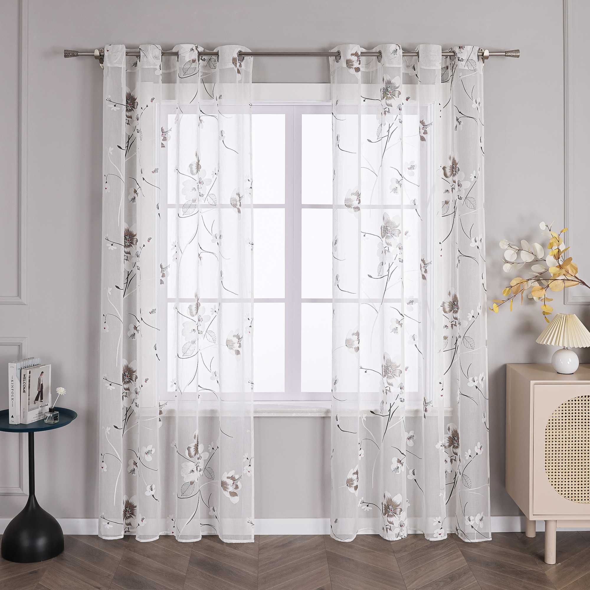 Das Bild zeigt ein Fenster, an dem ein Paar weiße Gardinen hängt. Die Gardinen sind mit einem filigranen Blumenmuster verziert, wobei die Blüten hauptsächlich in Weiß und Braun gehalten sind. Die Blütenmitte ist mit roten Akzenten verziert, und die Zweige sind in schwarzen Linien dargestellt. Die Gardinen sind an einer Metallstange befestigt, die an beiden Enden an einer hellgrauen Wand montiert ist.