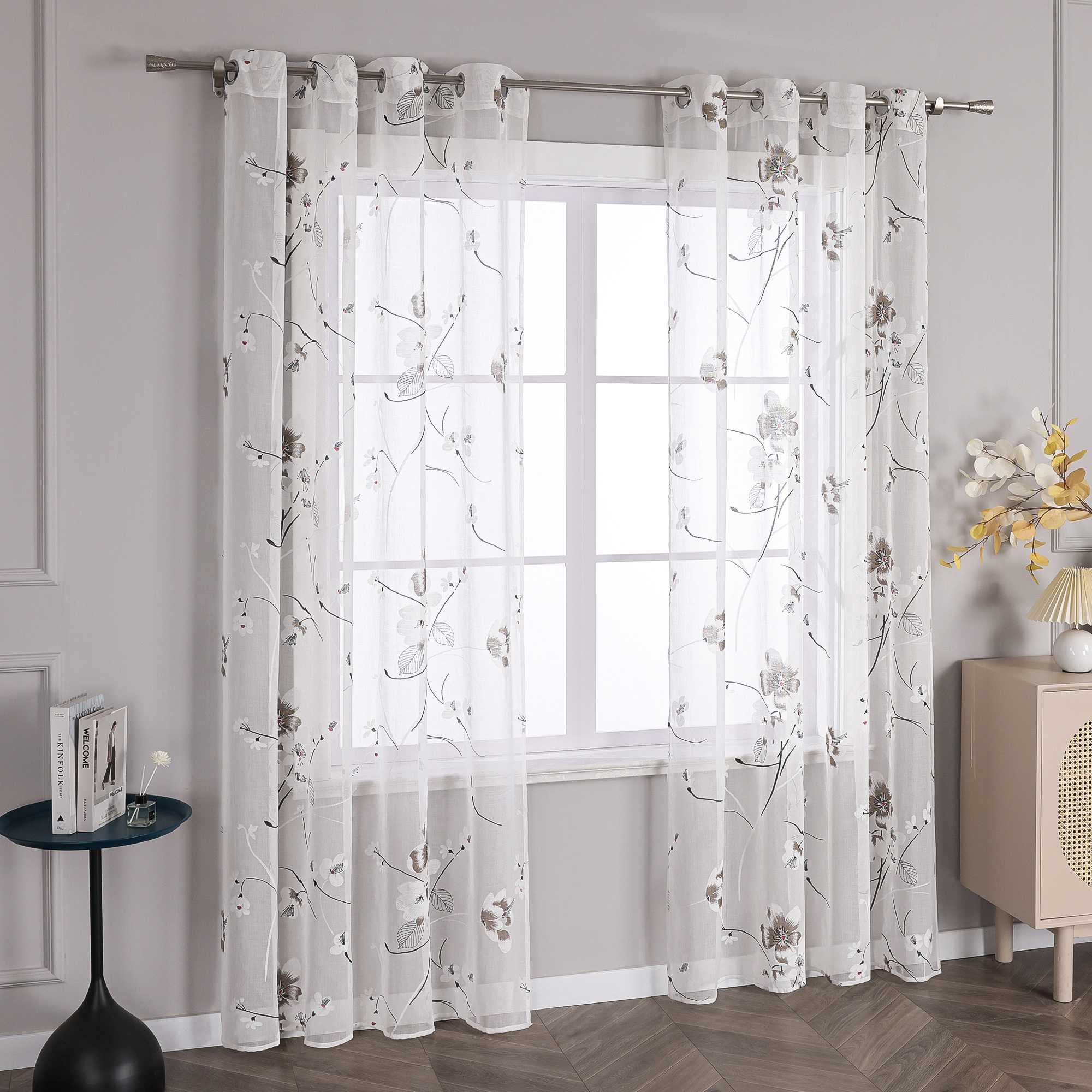 Dies ist eine Gardine mit Blumenmuster. Der Stoff ist leicht und durchsichtig, mit einem weißen Hintergrund, auf dem braune und schwarze Linien die Blätter und Zweige der Blumen in einem eleganten Design nachzeichnen.