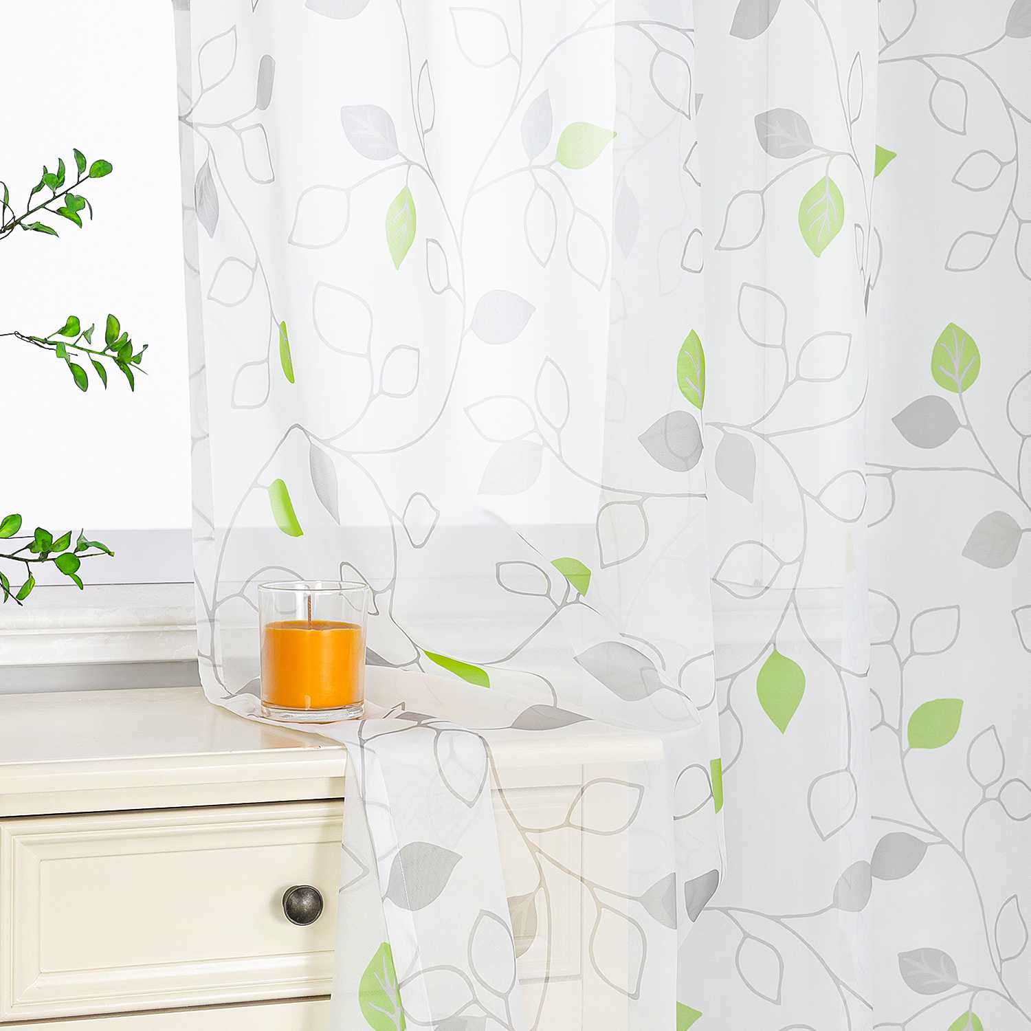 Das Bild zeigt weiße, transparente Vorhänge mit grünen und grauen Blättermustern. Auf dem weißen Schrank neben den Vorhängen steht eine orangefarbene Kerze, umgeben von grünen Pflanzen. Der Stil der Vorhänge ist frisch und natürlich, mit guter Lichtdurchlässigkeit, und schafft eine gemütliche Atmosphäre im Raum. Sie eignen sich besonders gut für die Fensterdekoration im Wohnzimmer oder Schlafzimmer.