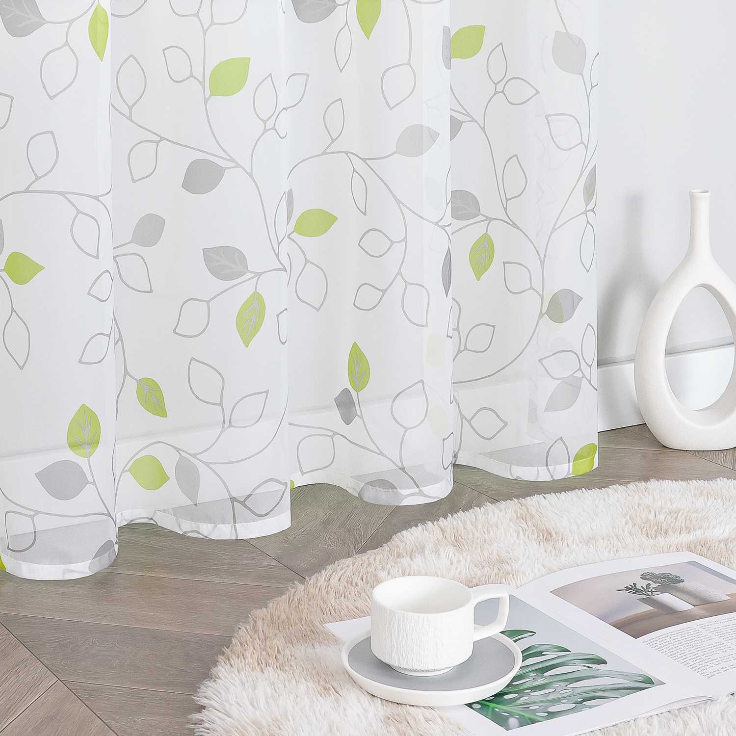 Dieser weiße Voile-Vorhang mit grünen und grauen Blattmustern ist leicht und lässt viel Licht durch. Neben dem Vorhang steht eine weiße Vase, auf dem weichen Teppich liegen eine Tasse mit Untertasse und ein aufgeklapptes Magazin. Der Vorhang schafft eine natürliche, frische Atmosphäre und eignet sich hervorragend für Wohnzimmer oder Schlafzimmer, um eine gemütliche und einladende Wohnumgebung zu gestalten.